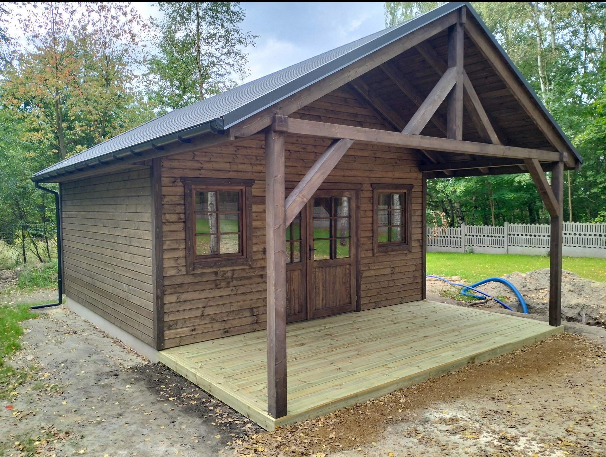 Drewniany domek z tarasem, elewacja z brązowych desek, okna z ciemnymi ramami, szary dach, zielone drzewa w tle, niebieski wąż na ziemi obok tarasu.