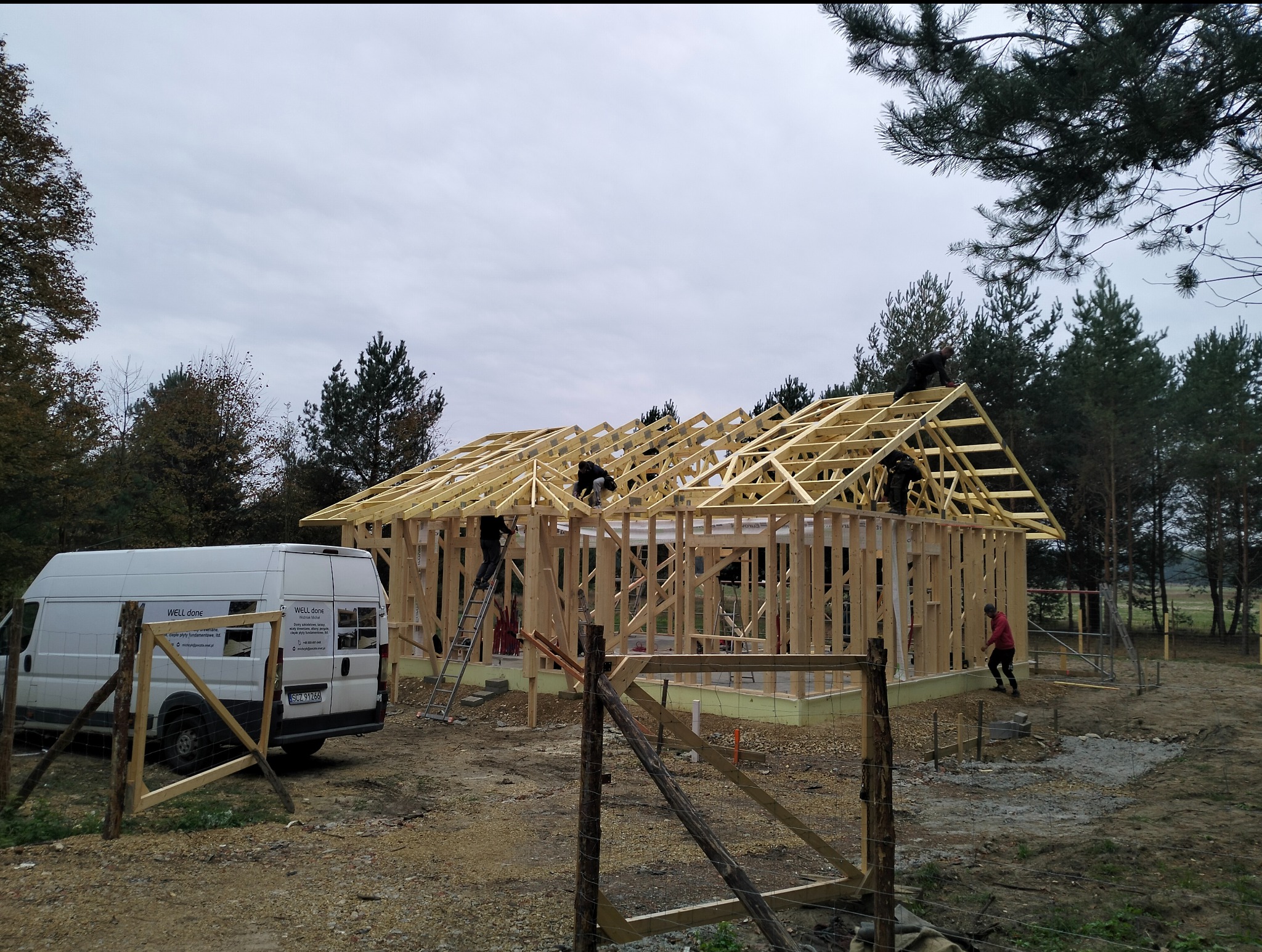 Konstrukcja drewniana domu szkieletowego w trakcie budowy, widoczni pracownicy na dachu i drabina. Na pierwszym planie prowizoryczne ogrodzenie, w tle las i bus firmy.