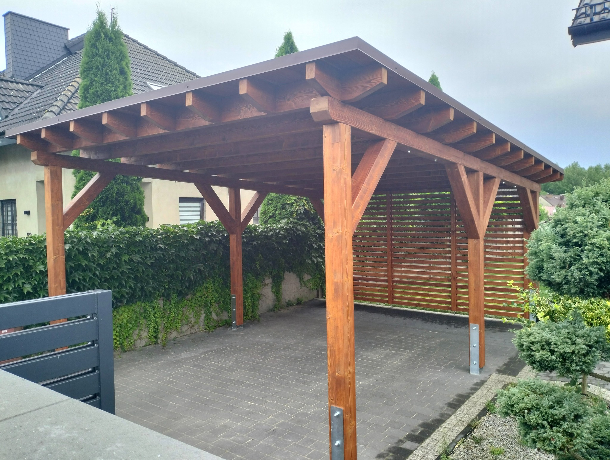 Drewniana wiata garażowa z brązowym zadaszeniem, wsparta na solidnych słupach, na tle zieleni i kostki brukowej. Widoczne detale konstrukcyjne.