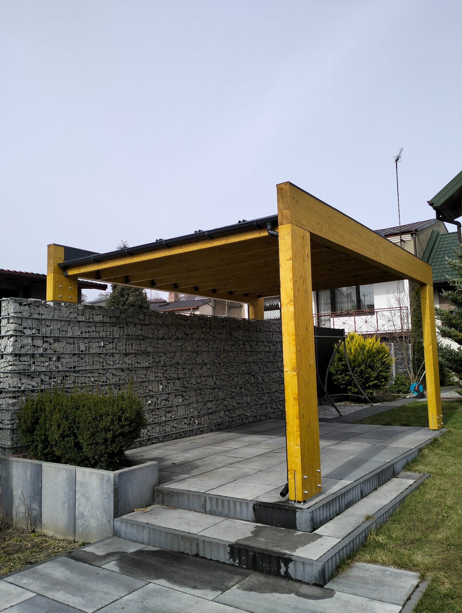Żółta drewniana pergola z czarnym dachem na kamiennej podłodze, obok gabionowego muru. Widok z zewnątrz, w tle dom z białymi ścianami i zielenią.