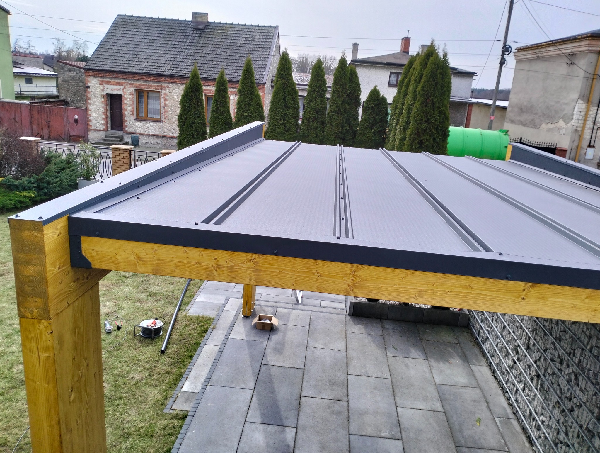 Drewniana pergola z szarym zadaszeniem na kamiennej podłodze, widok z góry. W tle domy i tuje. Widoczne narzędzia montażowe.
