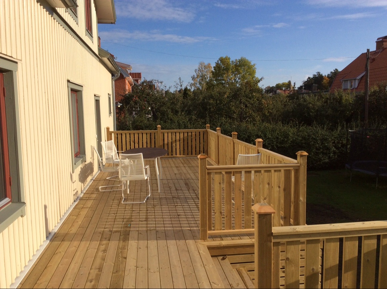 Drewniany taras przy domu szkieletowym z jasną elewacją, wyposażony w stół i krzesła. Widok na zieleń i zabudowę w tle, błękitne niebo.