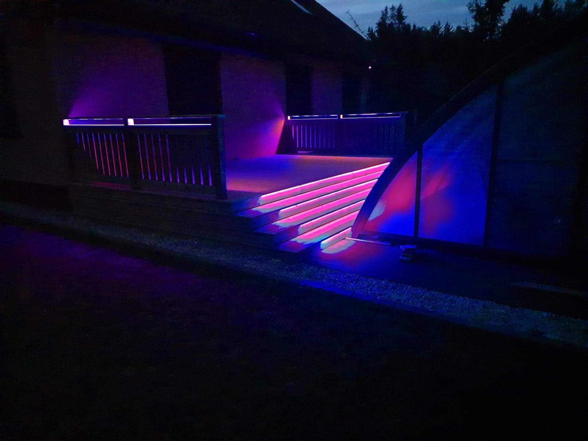 Oświetlony, drewniany taras domu szkieletowego nocą. Fioletowe i niebieskie światło LED podkreśla schody i balustrady, tworząc nowoczesny wygląd.