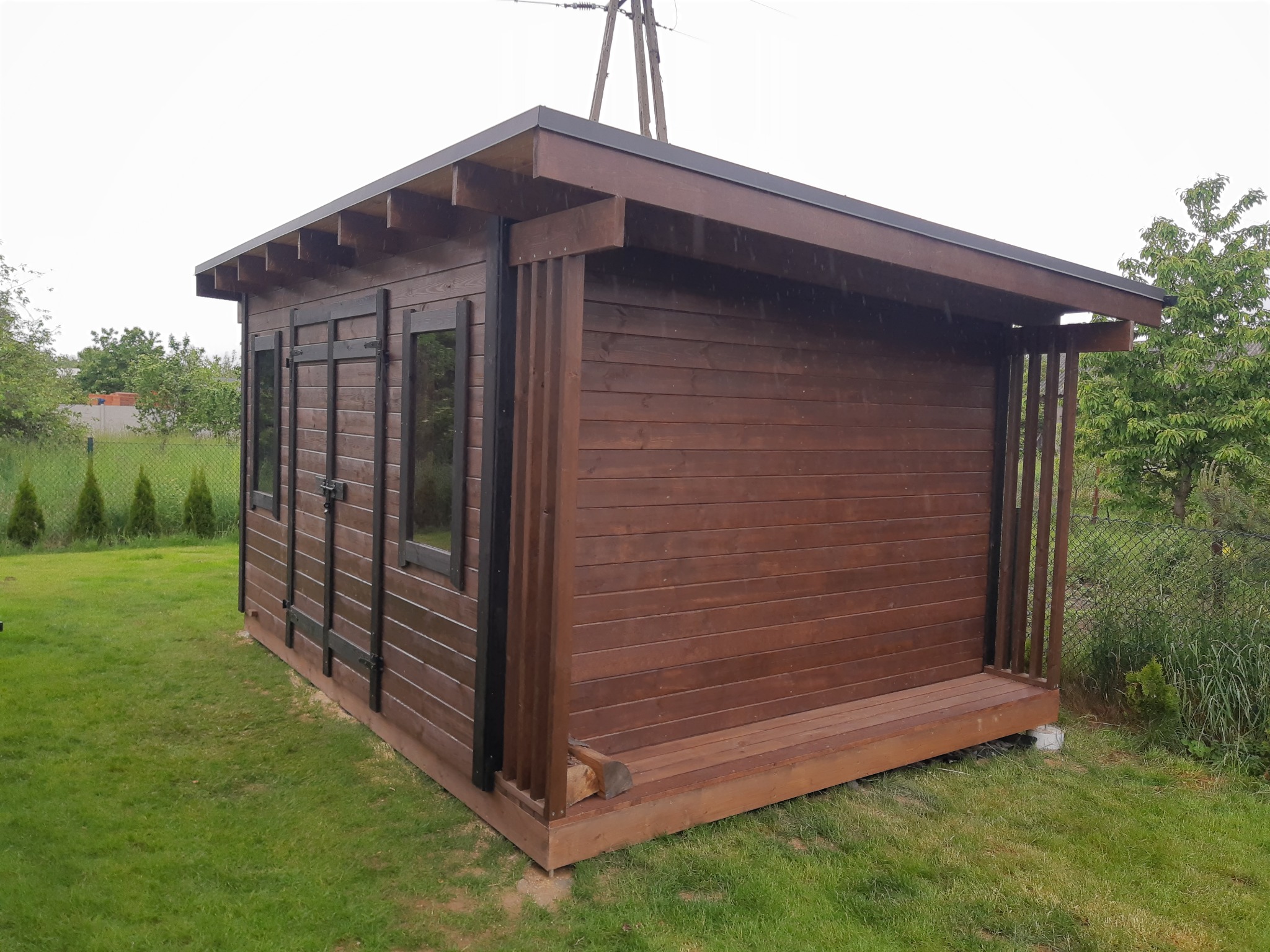 Gotowy, brązowy domek narzędziowy z drewna na trawie, z czarnymi okuciami okiennymi i małym tarasem. Widok z boku, w tle zieleń i słupy energetyczne.