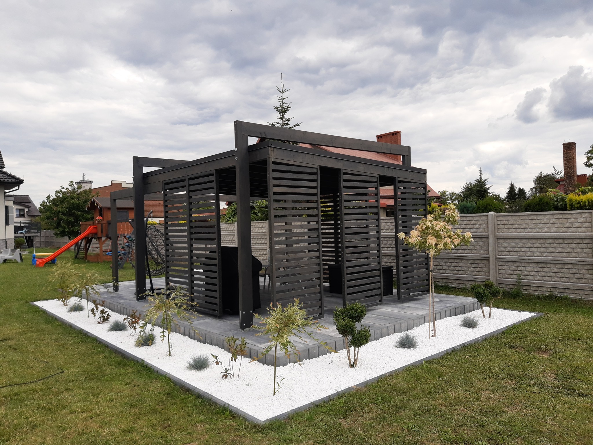 Nowoczesna, ciemna altana szkieletowa z poziomymi listwami, otoczona białymi kamieniami i zielenią, na tle trawnika i domów jednorodzinnych.