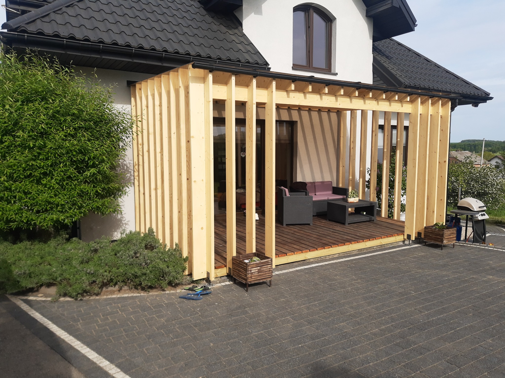 Drewniana pergola z meblami ogrodowymi przy domu z ciemną dachówką, widziana z zewnątrz, na tle zieleni. Widoczne klapki obok donicy.