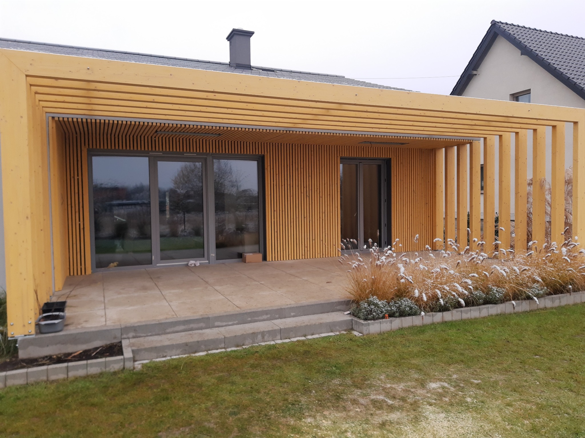 Nowoczesny dom szkieletowy z drewnianą pergolą i tarasem. Widoczne duże przesuwne okna i dekoracyjne trawy. Tło: inny dom i pochmurne niebo.