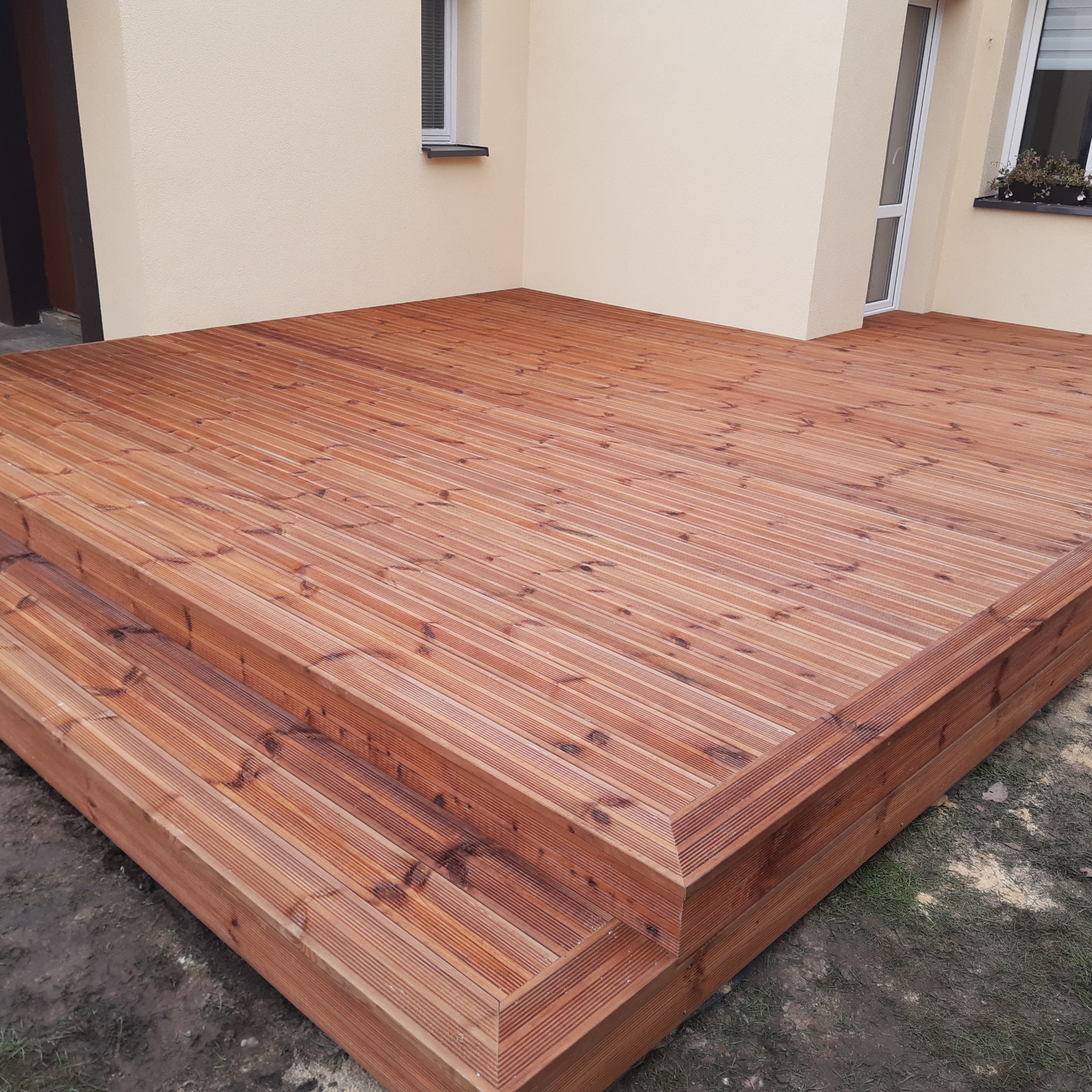 Drewniany taras z dwoma stopniami przy beżowym budynku z oknami i kwiatami. Deski ułożone równolegle, tworząc geometryczny wzór. Widok z dołu, podkreślający strukturę drewna.