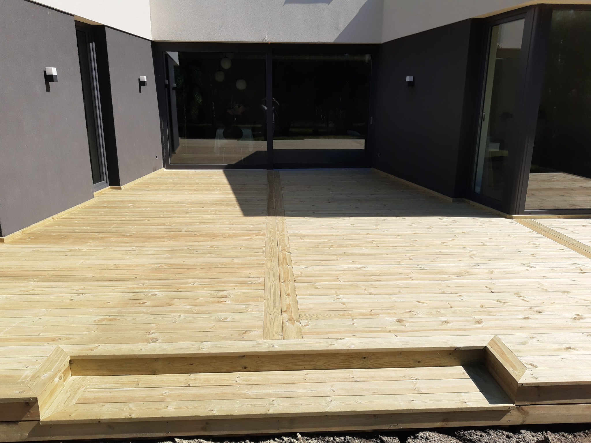 Nowoczesny taras z jasnego drewna przy domu z ciemnymi ścianami i dużymi oknami przesuwnymi. Schody tarasowe z drewna prowadzą na poziom gruntu.