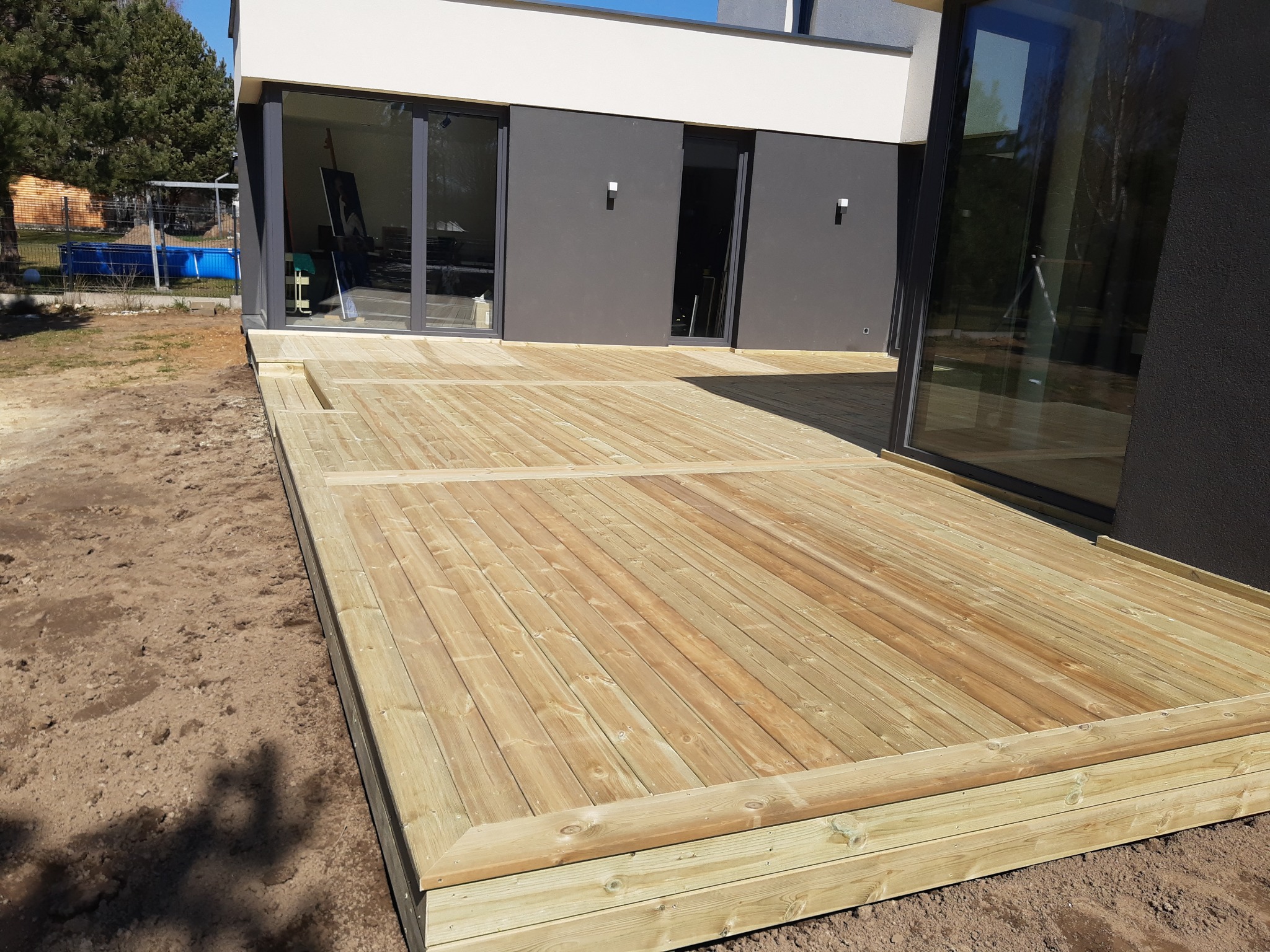 Nowoczesny taras z jasnego drewna przy domu szkieletowym z antracytową elewacją i dużymi oknami przesuwnymi. Widoczny fragment ogrodu z basenem.