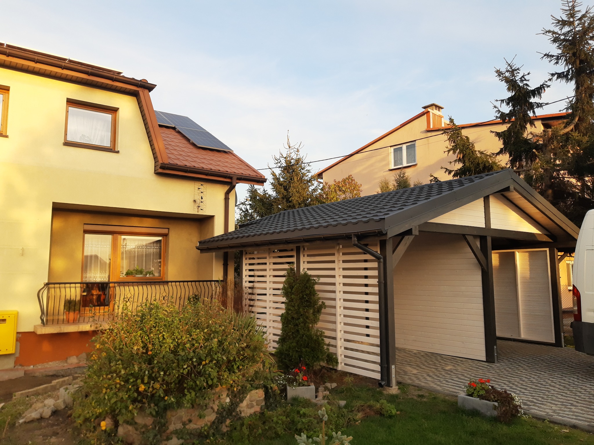 Żółty dom jednorodzinny z panelami słonecznymi na dachu i białą wiatą garażową z drewnianymi panelami, otoczony zielenią w słoneczny dzień.