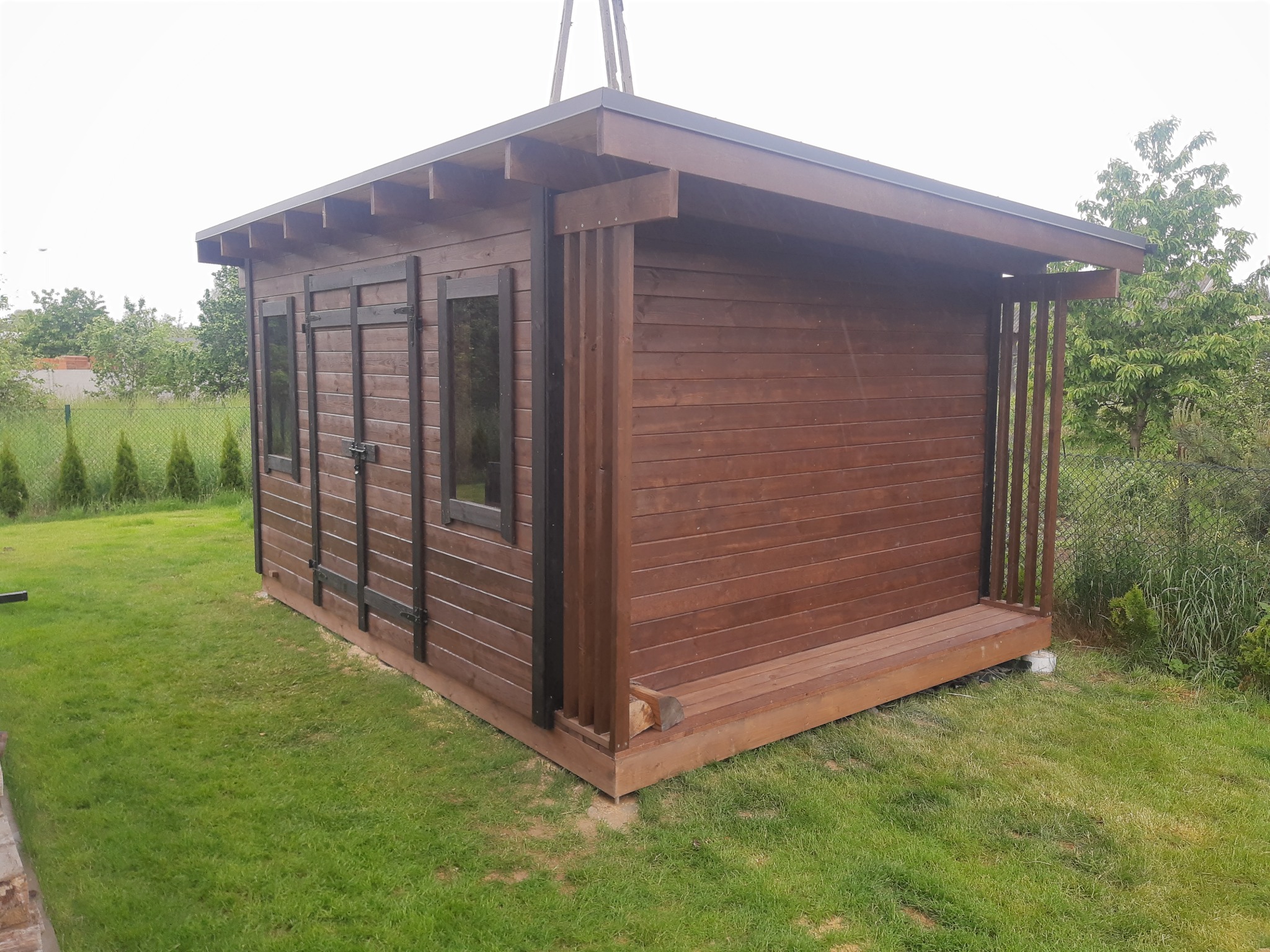 Drewniany domek narzędziowy z ciemnego drewna w Mstowie, z podestem i oknami, otoczony zielonym trawnikiem i krzewami. Widoczne fragmenty konstrukcji dachu.
