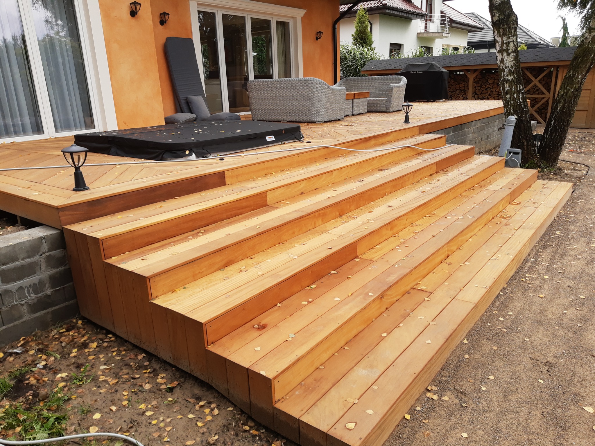 Drewniany taras z niskimi schodami, część domu z pomarańczową elewacją, meble ogrodowe i zadaszone miejsce na drewno w tle.