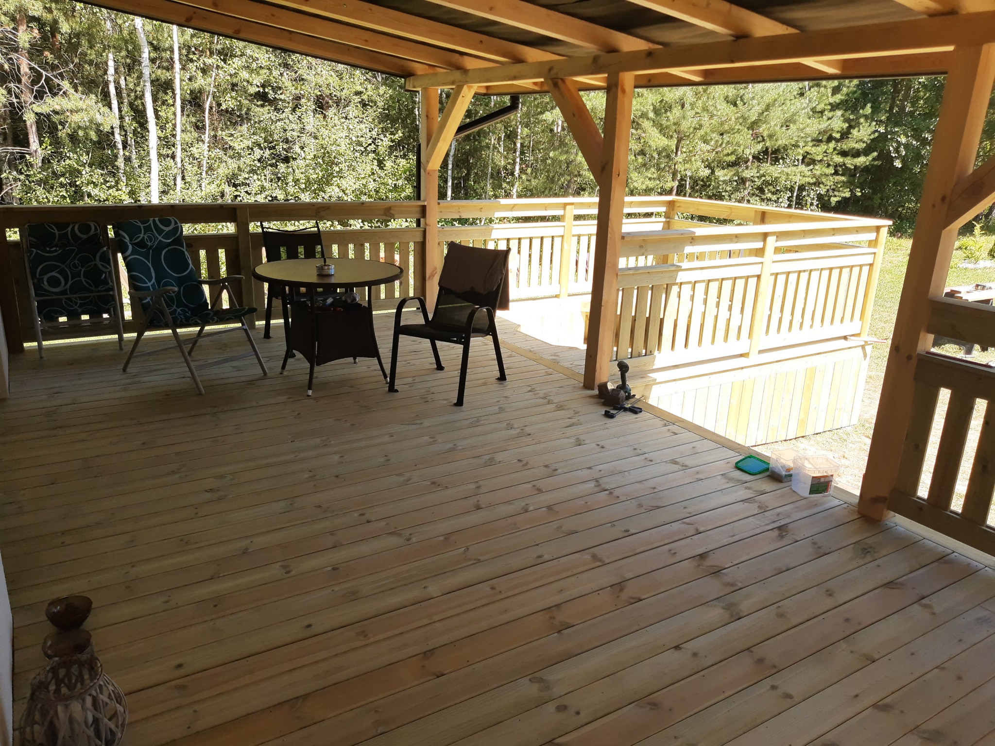 Drewniany taras z zadaszeniem, krzesłami i stołem, otoczony zielenią. Widoczne elementy konstrukcyjne z drewna i deski podłogowe. Częściowo zadaszony.