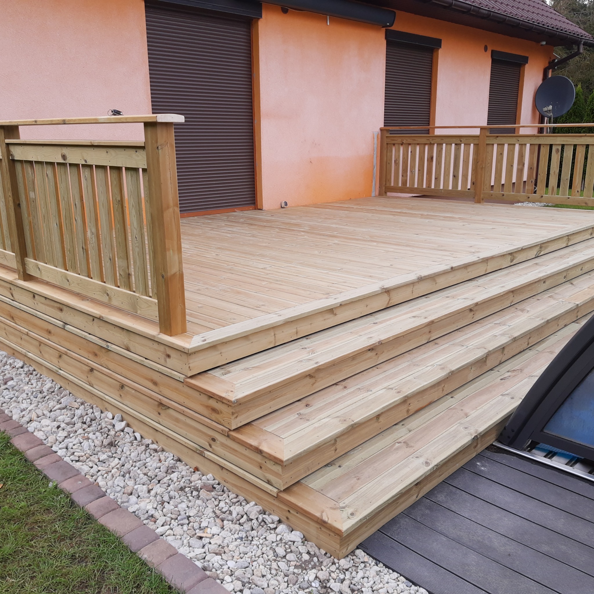 Drewniany taras z balustradą, schodami i kamiennym podsypem przy domu o pomarańczowej elewacji z brązowymi roletami. Widok z bliska na detale konstrukcji.