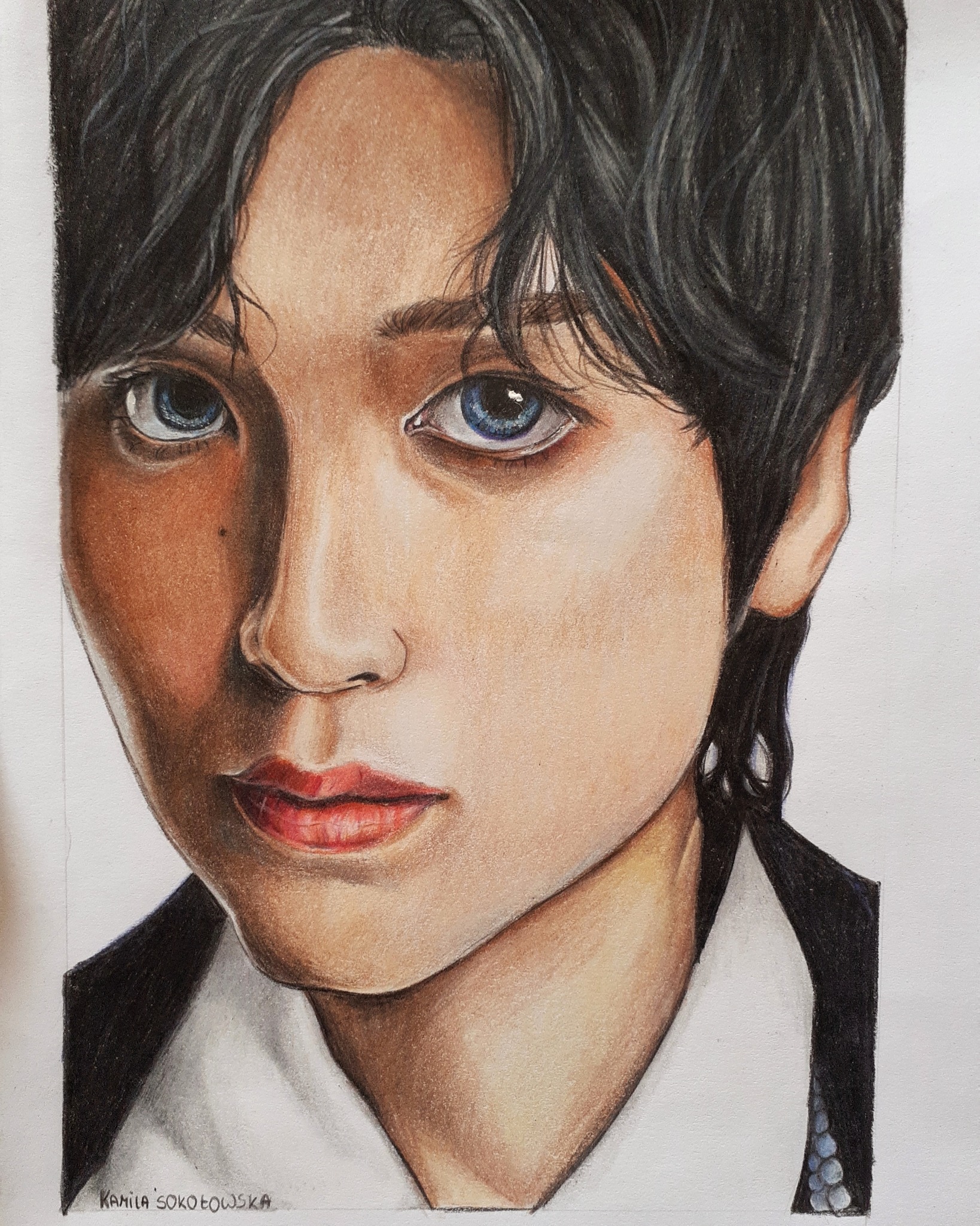 Portret młodej osoby z ciemnymi włosami i niebieskimi oczami, wykonany w technice rysunku kredkami, z widocznym cieniowaniem i detalami twarzy, podpisany 'Kamila Sokołowska'.