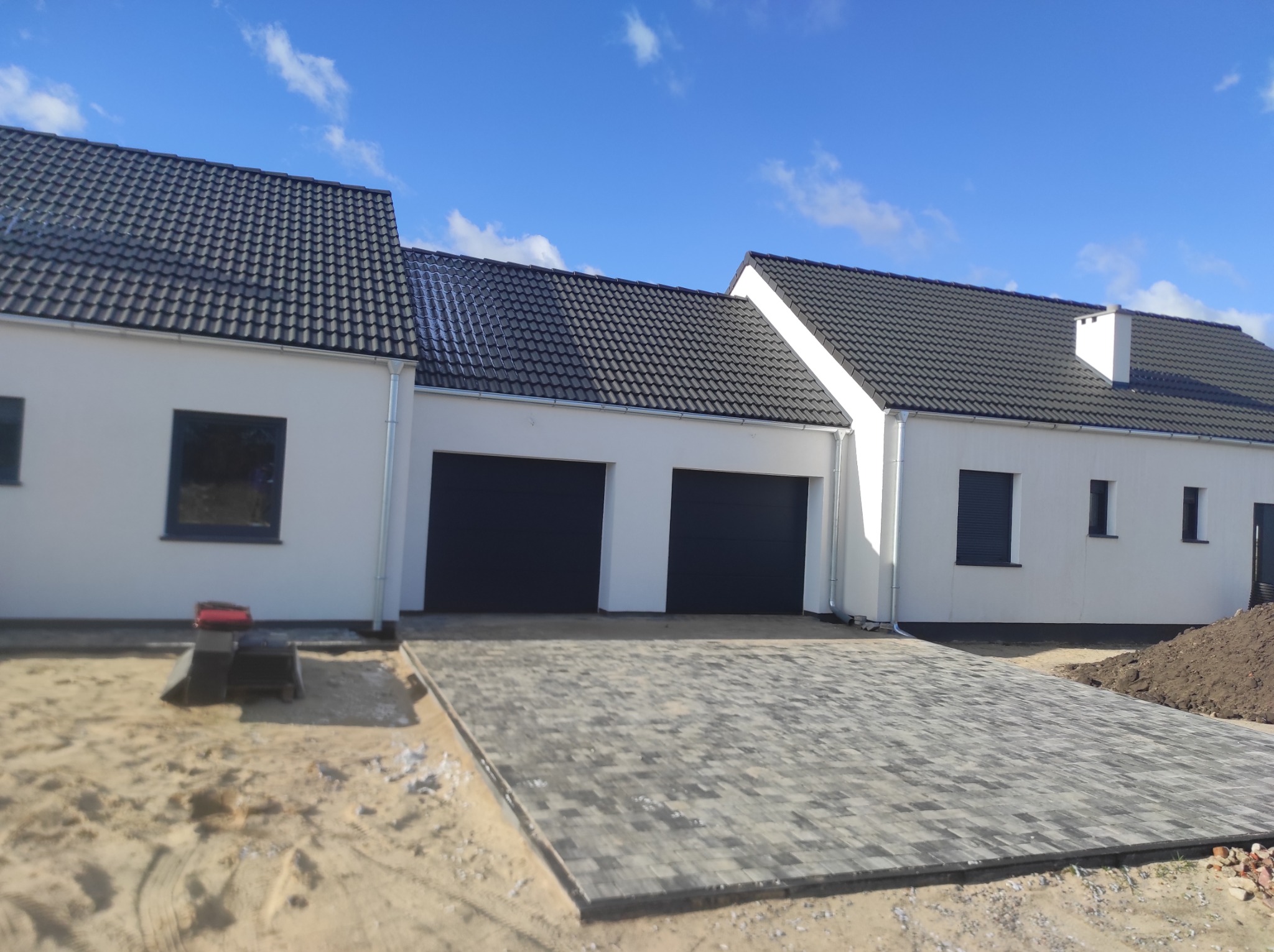 Nowo wybudowany dom jednorodzinny z dwustanowiskowym garażem i wybrukowanym podjazdem z kostki w odcieniach szarości, na tle błękitnego nieba z lekkimi chmurami.