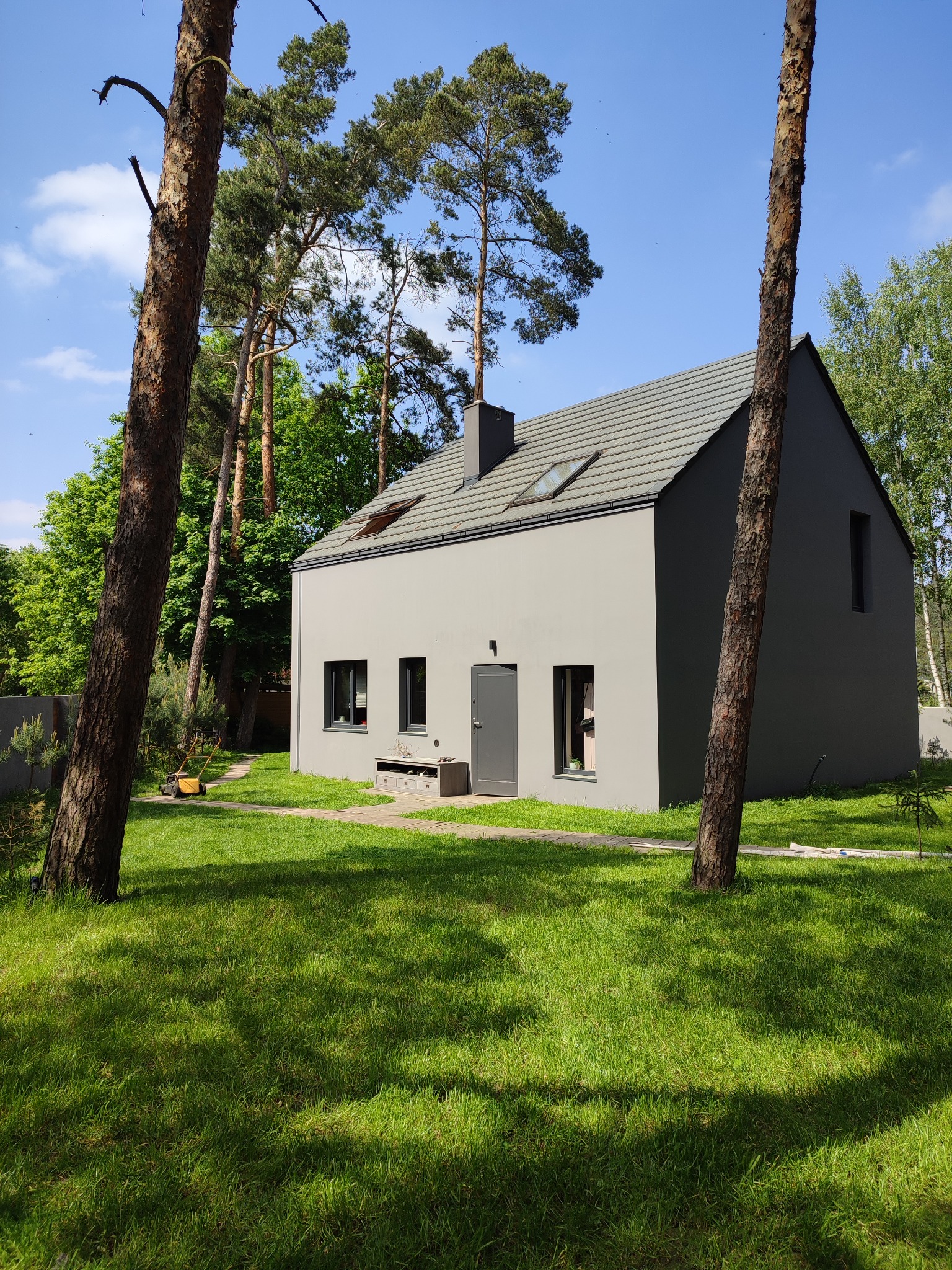 Nowoczesny dom jednorodzinny w kolorze szarości, z ciemną stolarką okienną i grafitowym dachem, otoczony zielonym trawnikiem i drzewami iglastymi w słoneczny dzień.