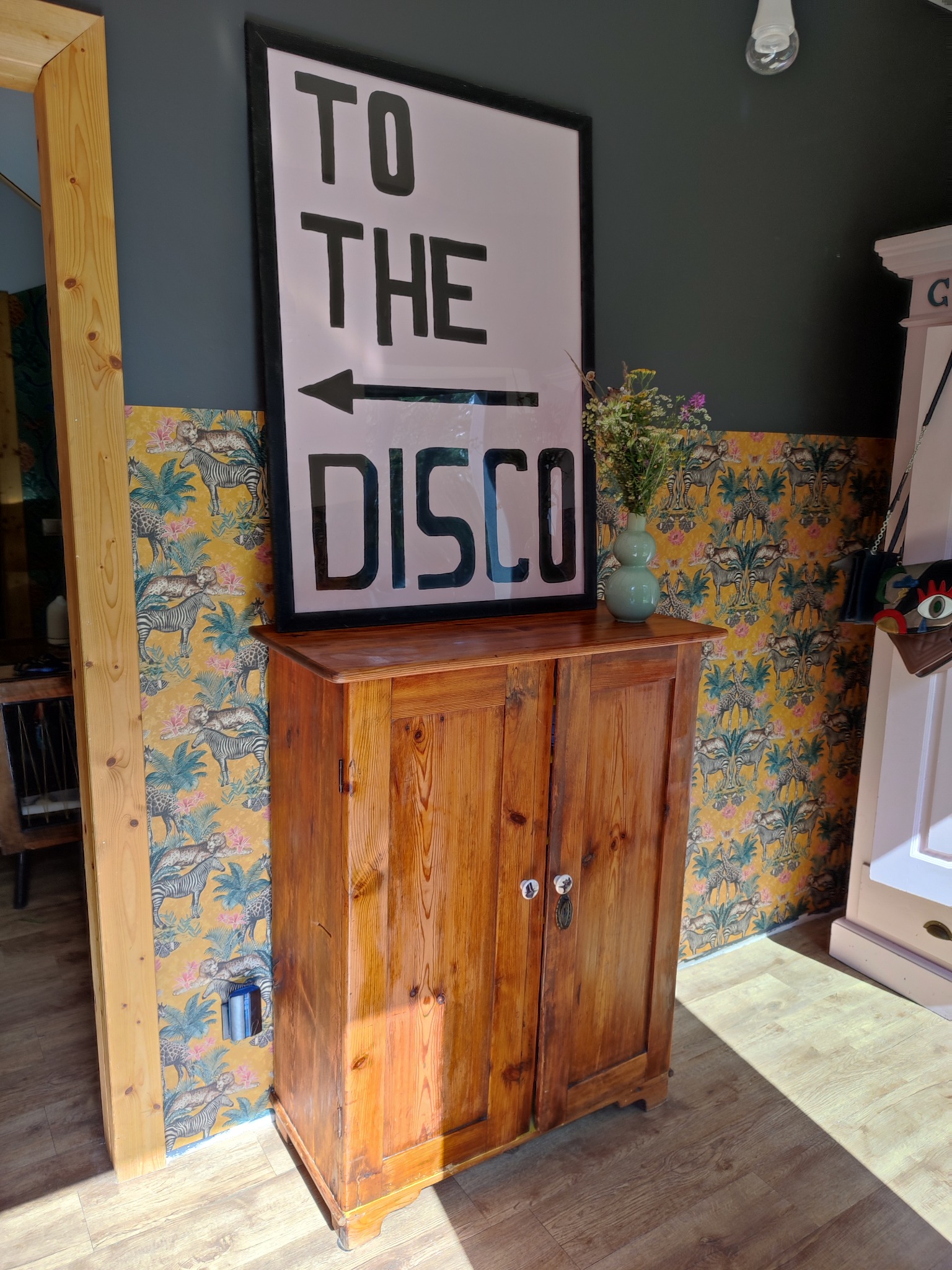 Drewniana szafka z plakatem 'TO THE DISCO' i wazonem z polnymi kwiatami na tle tapety w zwierzęce wzory.