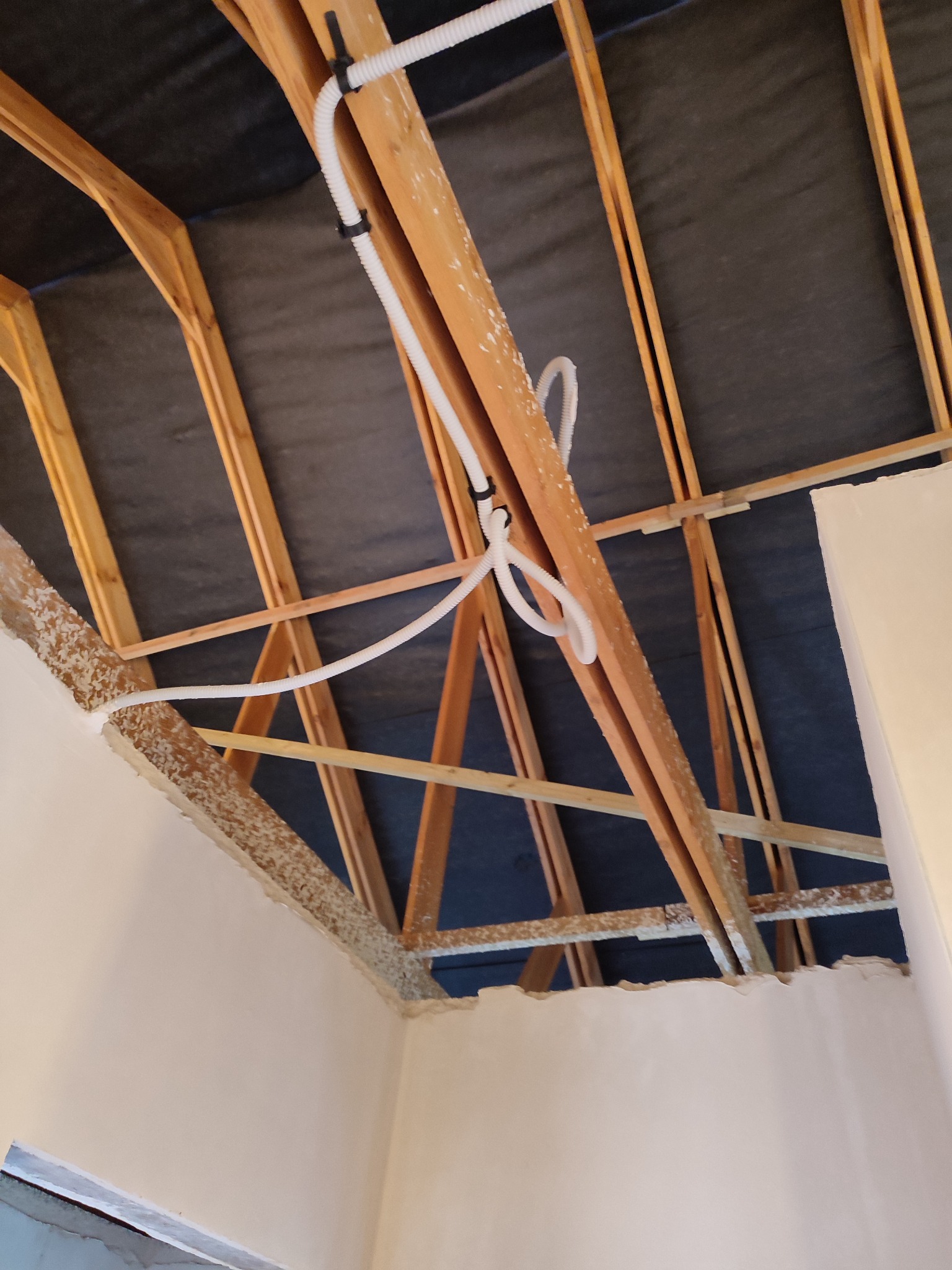 Surowy stan poddasza z widoczną konstrukcją drewnianą, izolacją oraz fragmentem ściany po nałożeniu gładzi. Wzdłuż krokwi poprowadzona biała, karbowana rurka instalacyjna przymocowana czarnymi...