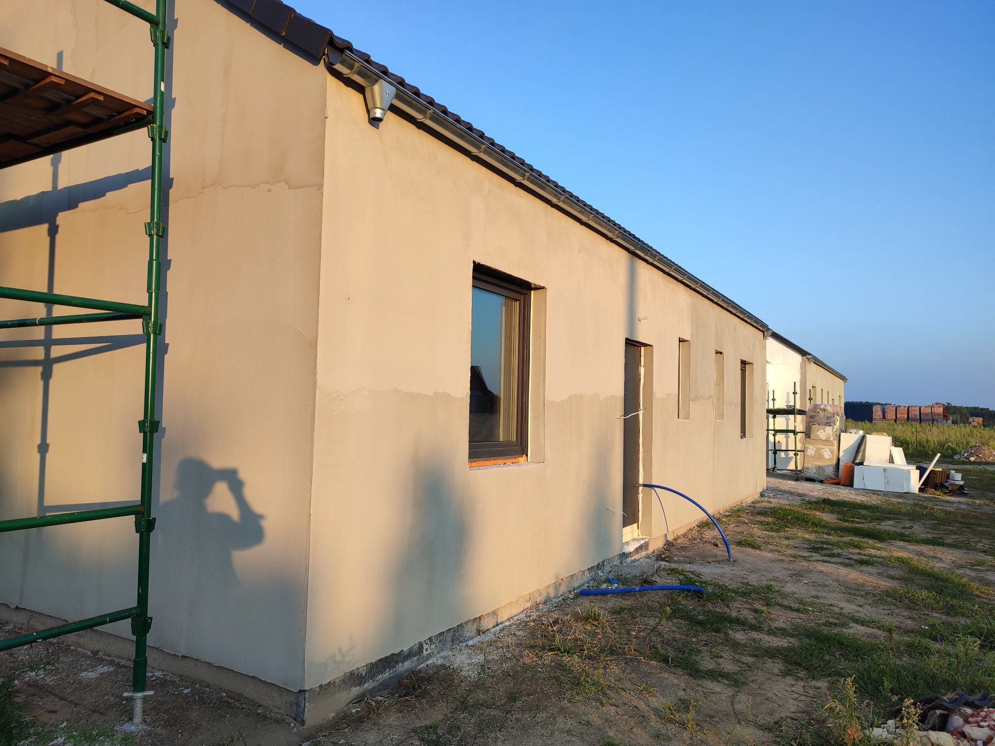 Świeżo otynkowana elewacja domu jednorodzinnego w trakcie prac wykończeniowych, widoczne rusztowanie, cień fotografa na ścianie, niebieski wąż i materiały budowlane w tle.