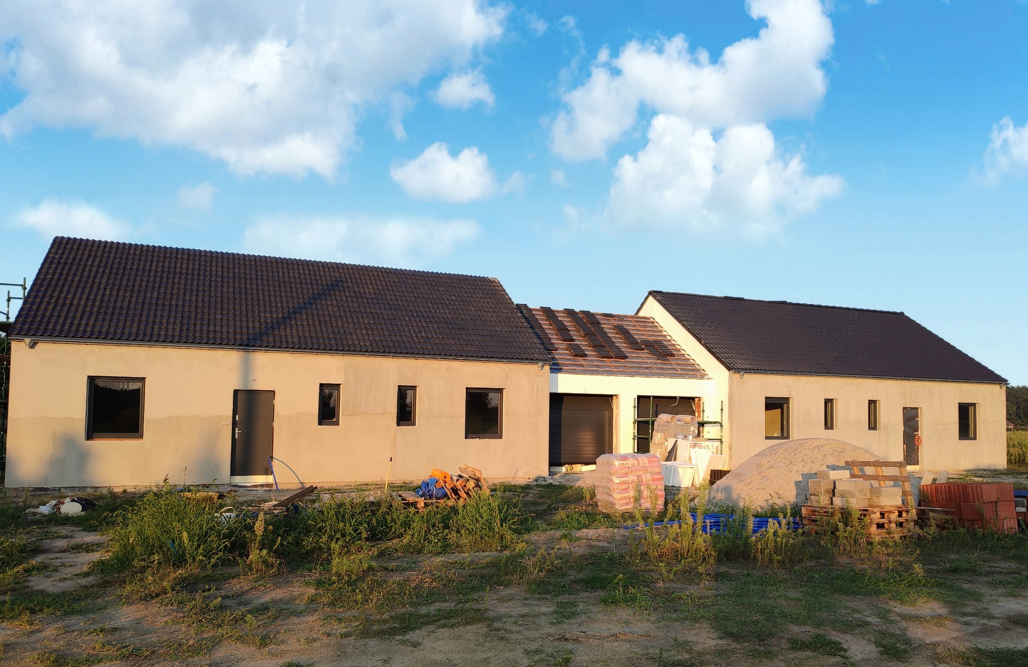 Niedokończony dom jednorodzinny w Śremie z widocznymi etapami prac: ocieplona elewacja, częściowo położony dach z czarnej dachówki, przygotowane materiały budowlane na placu budowy.