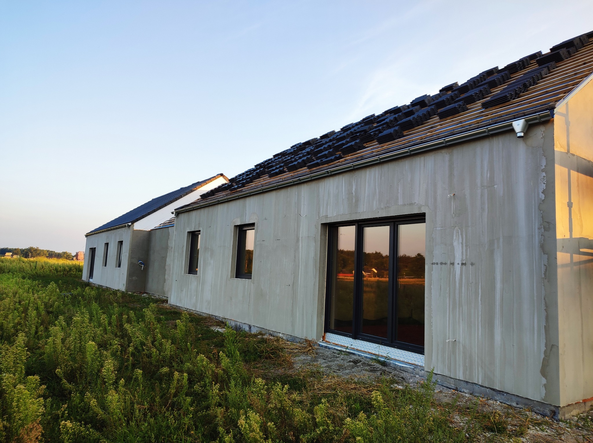 Surowa elewacja domu jednorodzinnego w trakcie prac, z widocznymi śladami po zaprawie i przygotowanym dachem z czarną dachówką, otoczona wysoką trawą i zielenią.