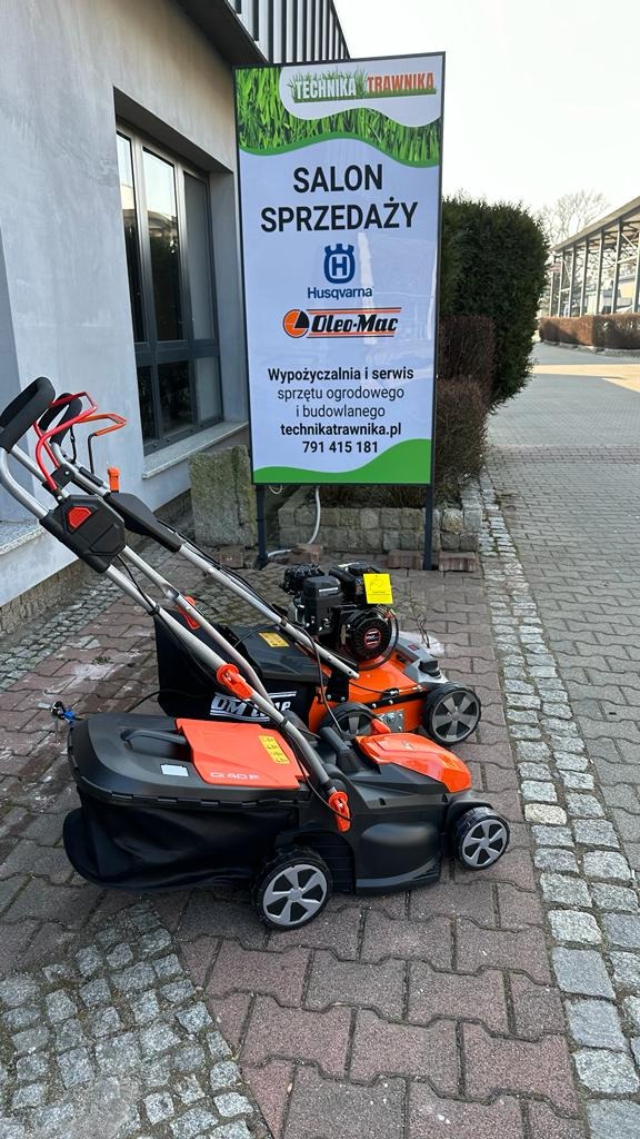 Dwie kosiarki spalinowe i elektryczne marki Husqvarna i Oleo-Mac przed salonem sprzedaży sprzętu ogrodowego Technika Trawnika z widocznym szyldem reklamowym.