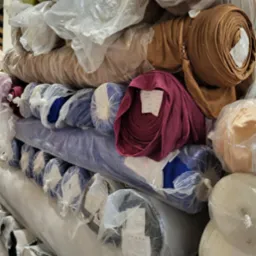 Magazyn rolek tkanin w folii, widoczne kolory: brązowy, fioletowy, niebieski i biały. Różne faktury i grubości materiałów.