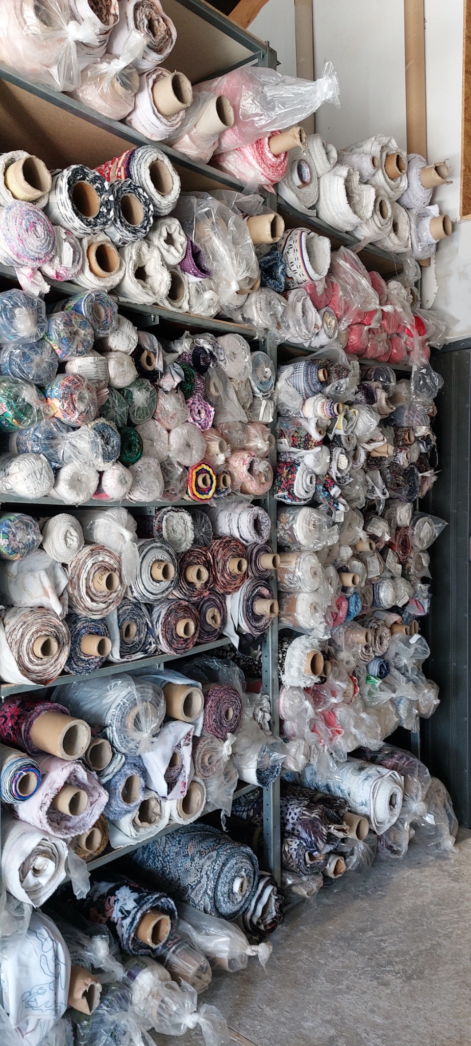 Regał magazynowy wypełniony rolkami różnorodnych tkanin, niektóre zabezpieczone folią, ustawione ciasno obok siebie.