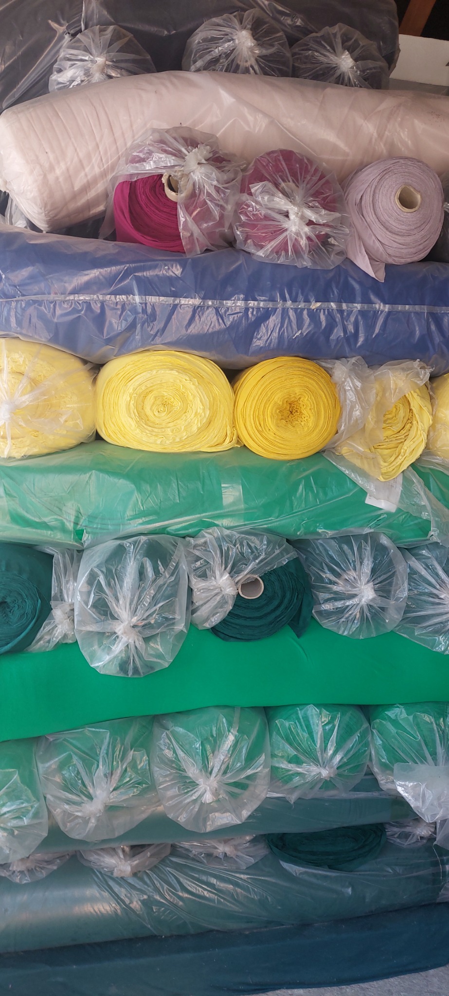 Stos rolek tkanin w różnych kolorach (różowy, fioletowy, niebieski, żółty, zielony), część z nich zapakowana w przezroczystą folię.