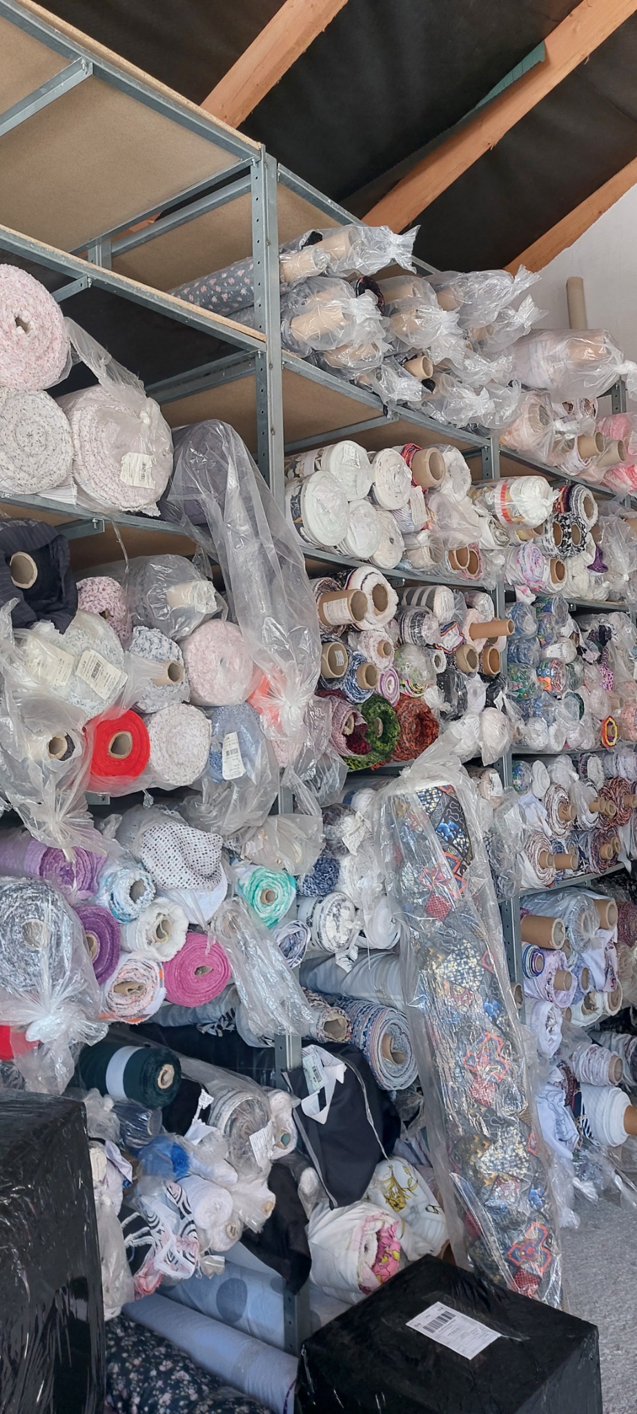 Magazyn z półkami wypełnionymi rolkami tkanin o różnych wzorach i kolorach, niektóre zafoliowane, widoczne drewniane elementy konstrukcji dachu.