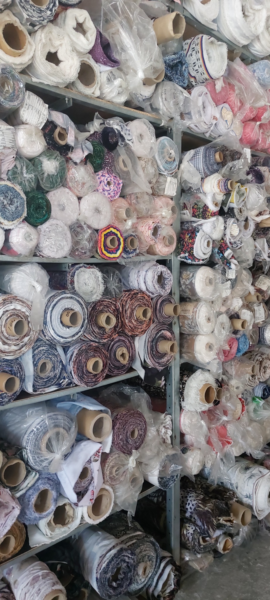 Pełne regały magazynowe z różnorodnymi rolkami tkanin, niektóre zapakowane w przezroczyste folie, widoczne wzory i tekstury materiałów.