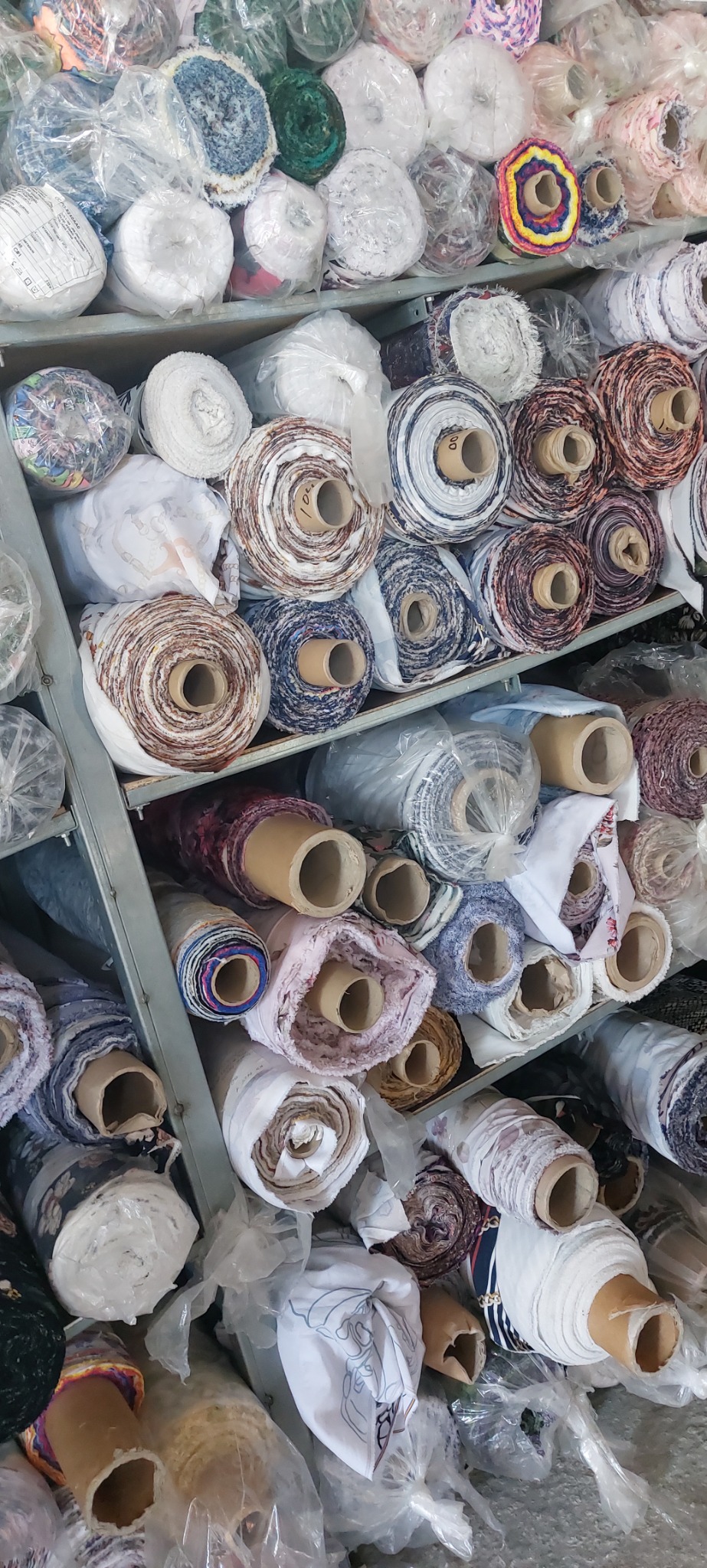 Półki w hurtowni tkanin wypełnione różnorodnymi rolkami materiałów, niektóre zapakowane w folię ochronną, widoczne wzory i tekstury tkanin.