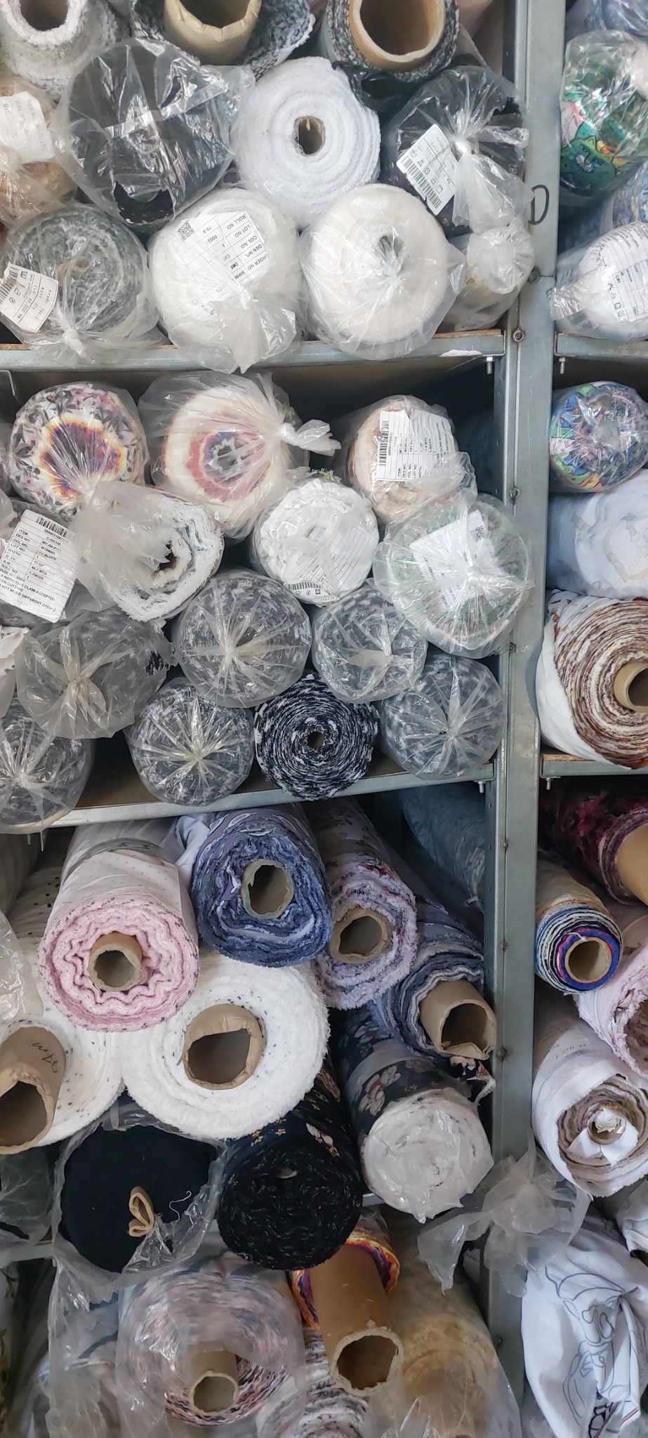 Półki regału wypełnione różnorodnymi rolkami tkanin, niektóre zapakowane w folię, widoczne etykiety z numerami partii i specyfikacją materiałów.