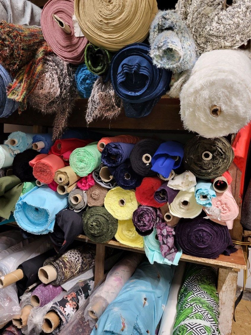 Półki regału wypełnione różnorodnymi rolkami tkanin o różnych fakturach, kolorach i wzorach, od gładkich po futerkowe, niektóre z widocznymi tekturowymi rdzeniami.