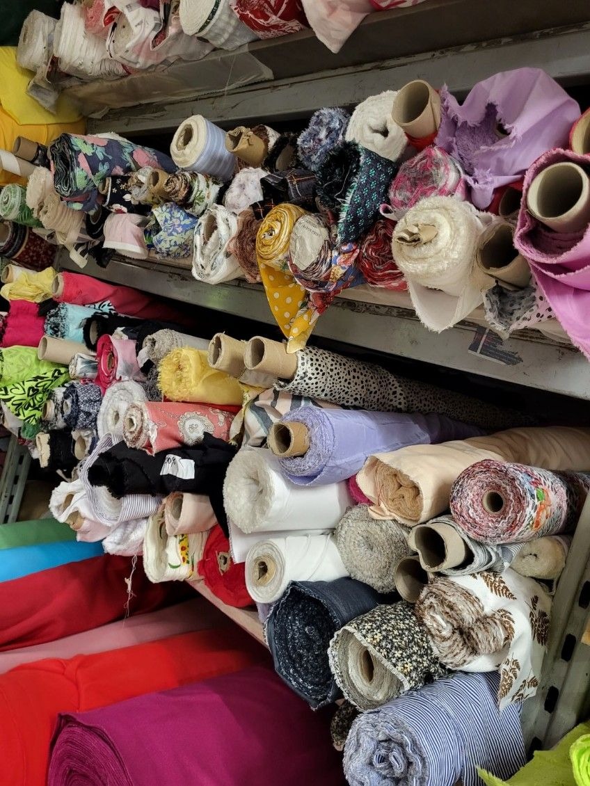 Regał magazynowy wypełniony różnorodnymi rolkami tkanin i dzianin, w tym wzorzyste, gładkie, kolorowe i białe, ułożone warstwami, widoczne tekturowe gilzy wewnątrz rolek.