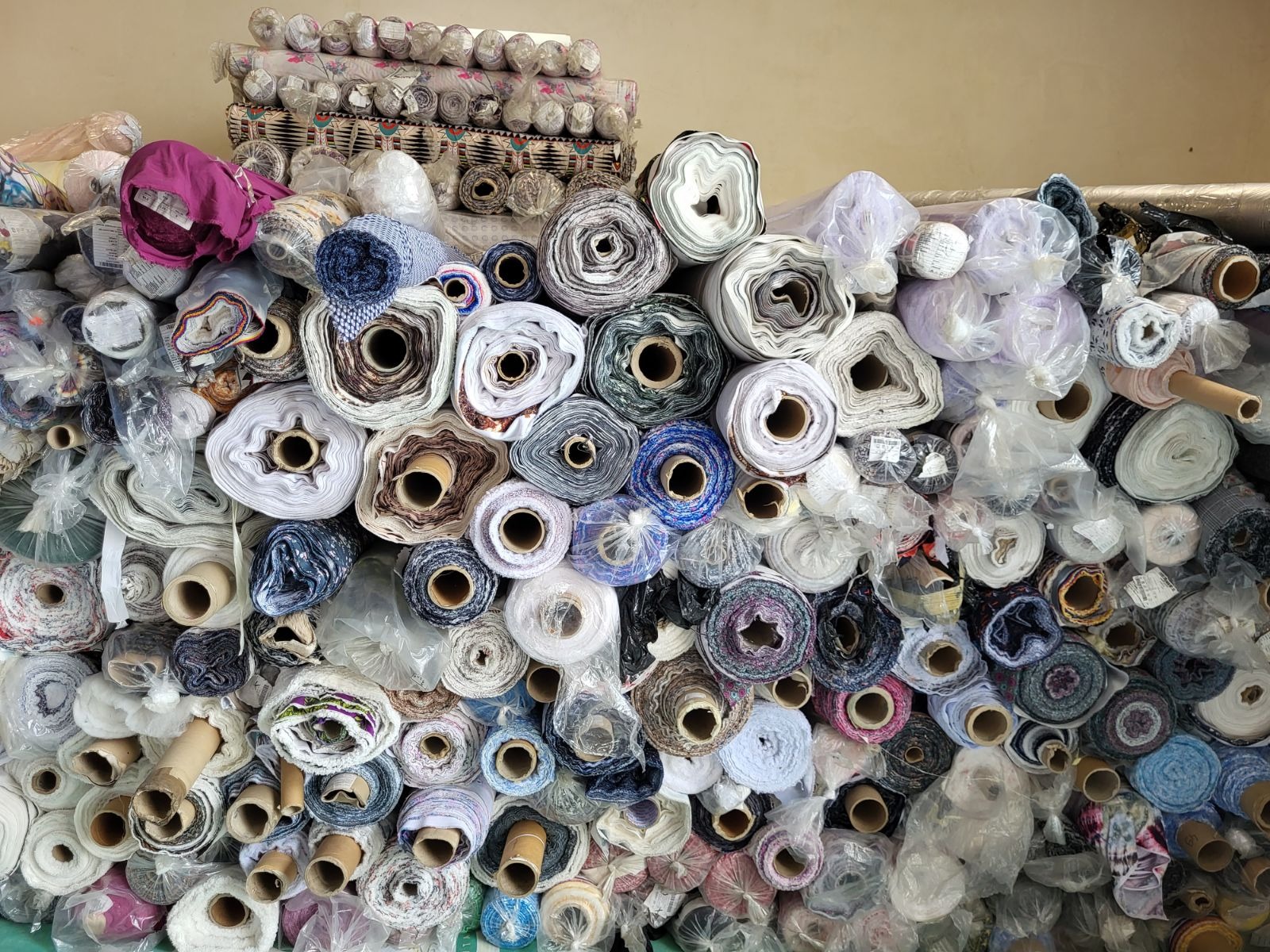 Stos różnorodnych tkanin w rolkach, niektóre zapakowane w folię, ułożone na sobie w hurtowni, widoczne różne faktury i kolory materiałów.