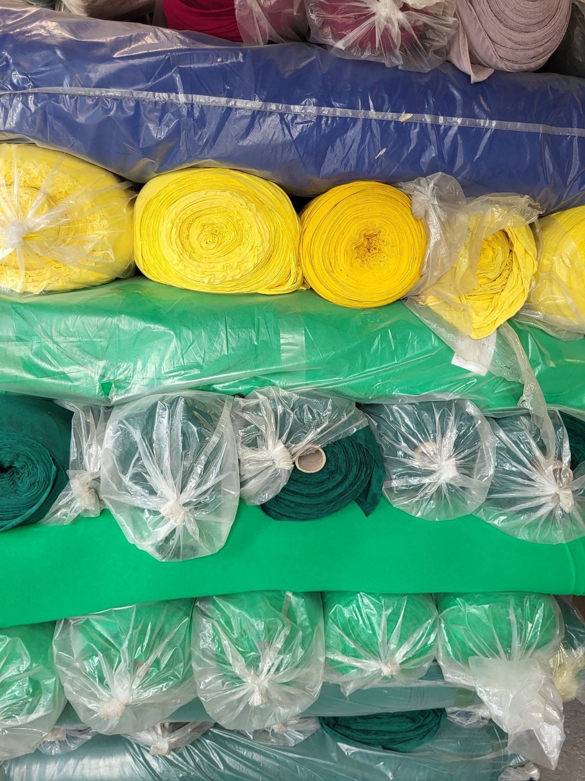Stos zwiniętych bel materiałów w różnych kolorach: niebieskim, żółtym i zielonym, niektóre zapakowane w przezroczyste folie.