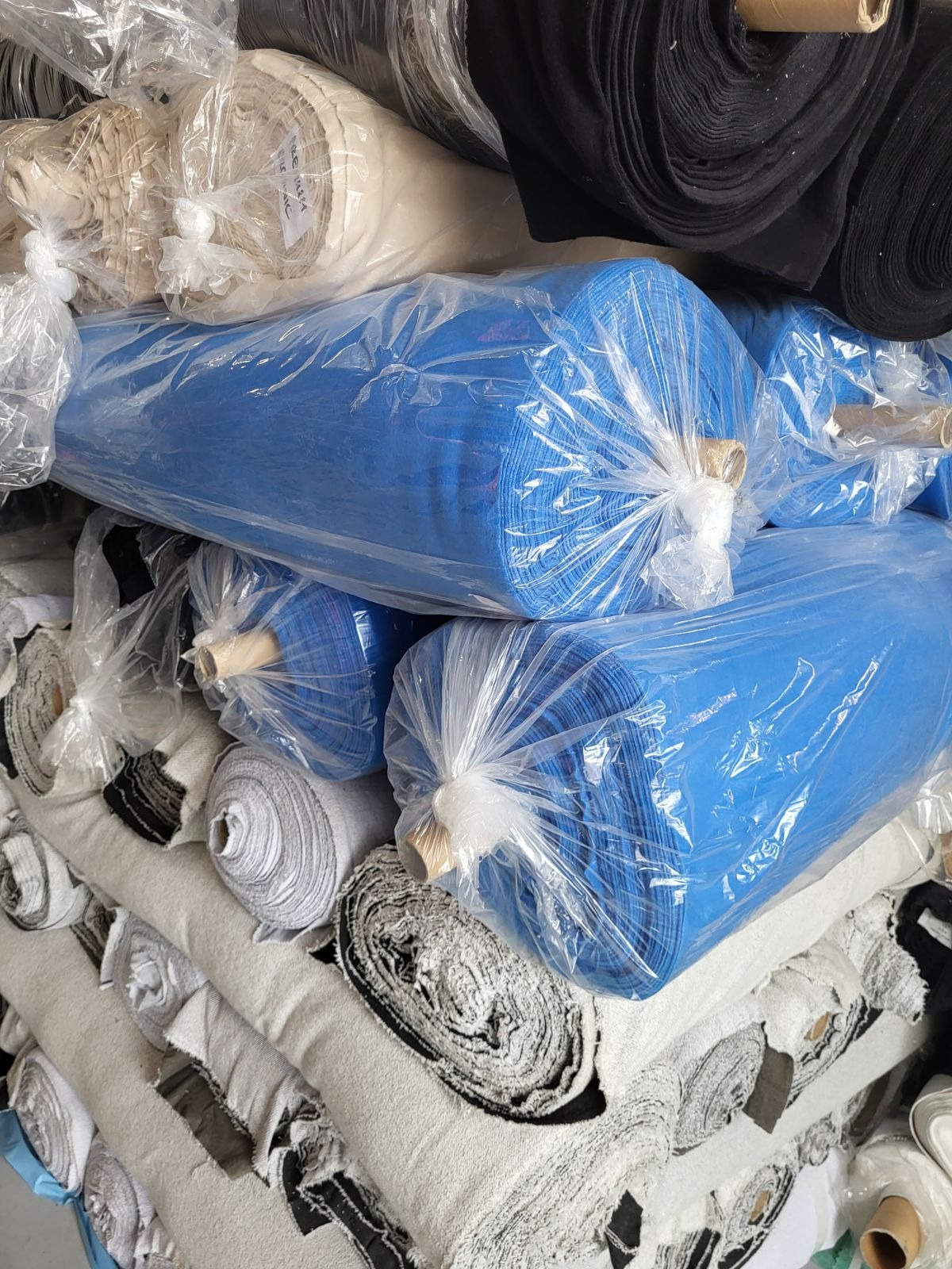 Stos rolek tkanin w różnych kolorach i wzorach, zapakowanych w folię, z widocznymi etykietami na niektórych rolkach.