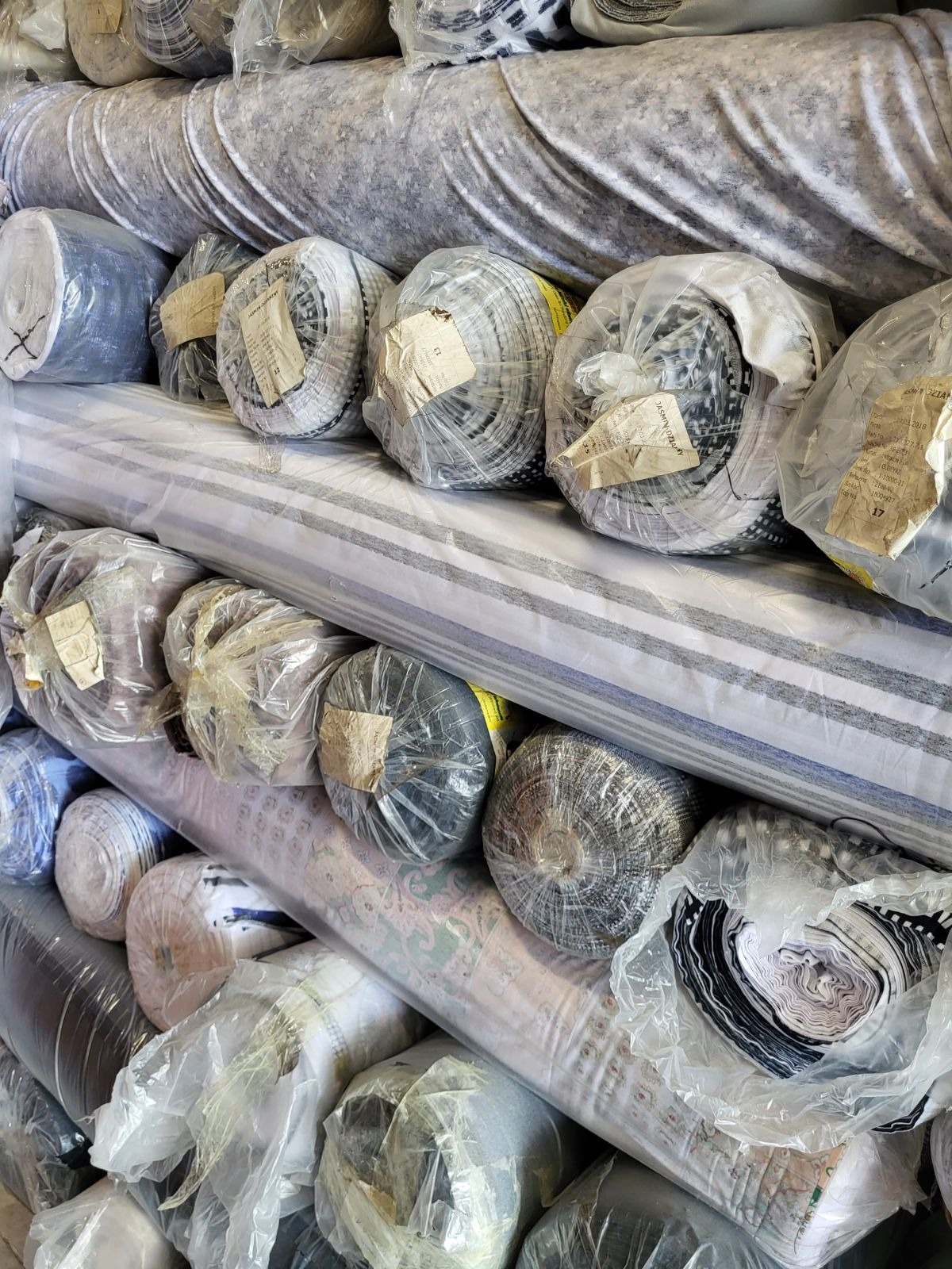 Stos rolek tkanin o różnorodnych wzorach i kolorach, owiniętych folią, z etykietami informacyjnymi, ułożonych warstwami w magazynie.