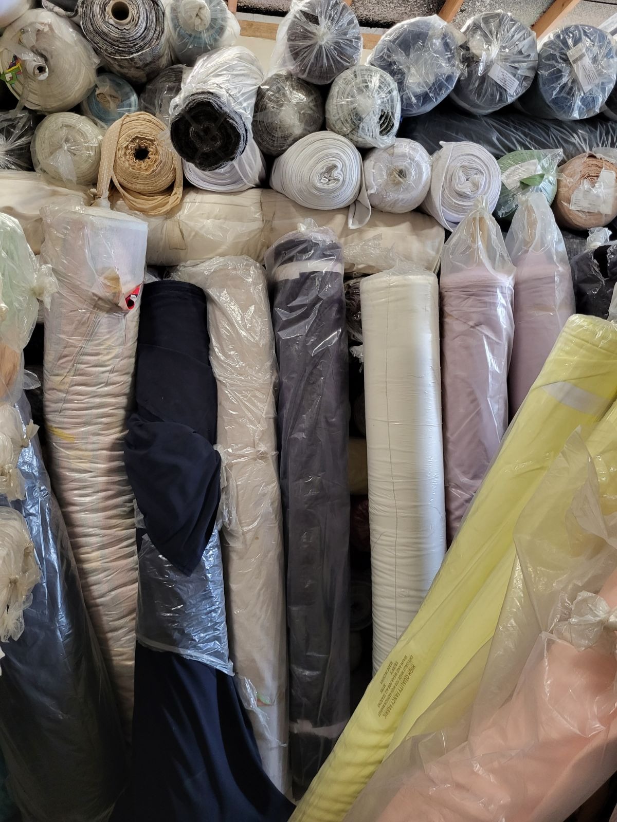 Magazyn rolek tkanin w różnych kolorach i fakturach, niektóre zapakowane w folię ochronną, ułożone pionowo i poziomo, widoczne także etykiety na niektórych rolkach.