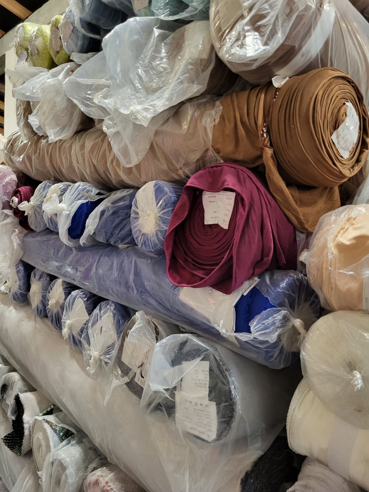 Magazyn rolek tkanin w folii, widoczne kolory: brązowy, fioletowy, niebieski i biały. Różne faktury i grubości materiałów.