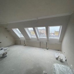 KondorBUD - Poddasze po szpachlowaniu, widoczne trzy okna dachowe z widokiem na miasto. Ściany przygotowane do malowania, na podłodze resztki materiałów budowlanych i narzędzia.