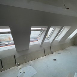 KondorBUD - Poddasze w trakcie prac wykończeniowych: widoczne okna dachowe, ściany przygotowane do malowania, surowa podłoga. Elementy montażowe na ścianach.
