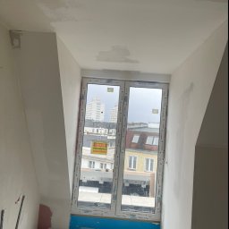 KondorBUD - Poddasze w trakcie wykańczania: okno z widokiem na miasto, ściany częściowo pokryte gładzią, widoczne niedoskonałości i przygotowania do dalszych prac wykończeniowych.