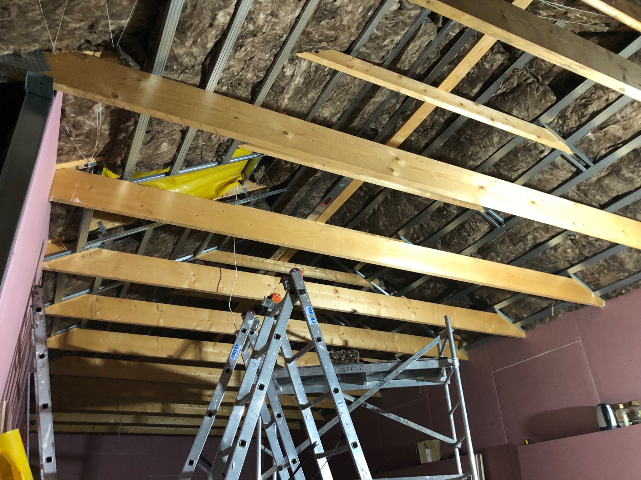 Skośny strych z izolacją, metalową konstrukcją i drewnianymi belkami, widok z dołu, drabina w tle. Prace wykończeniowe poddasza.