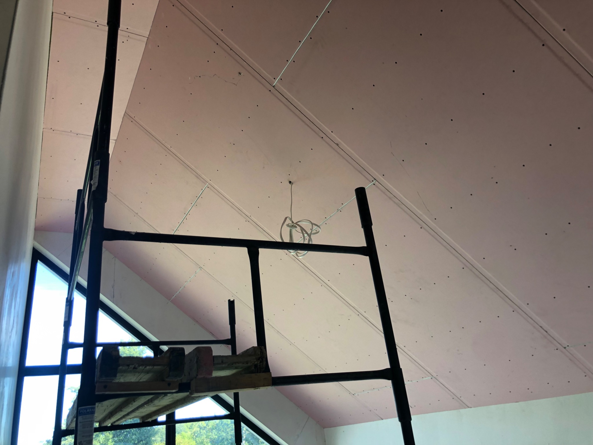 Skośny sufit poddasza z płyt kartonowo-gipsowych, widoczny ze stojącego rusztowania. Przewody elektryczne zwisają z sufitu. Okno trójkątne w tle.
