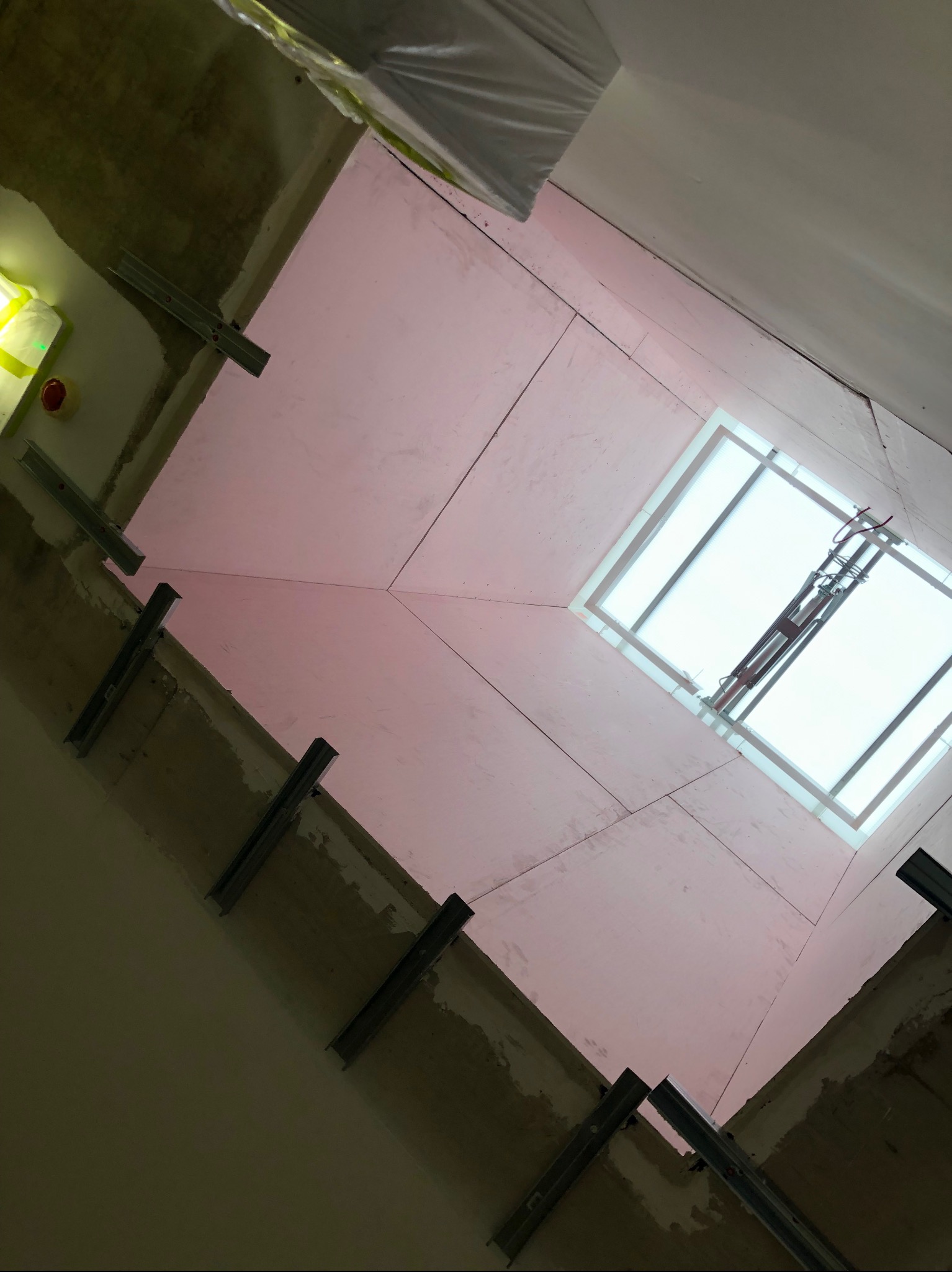 Rzut z dołu na montaż płyt karton-gips na suficie, widoczne metalowe profile i okno dachowe. Prace wykończeniowe w trakcie realizacji, sufit w kolorze delikatnego różu.