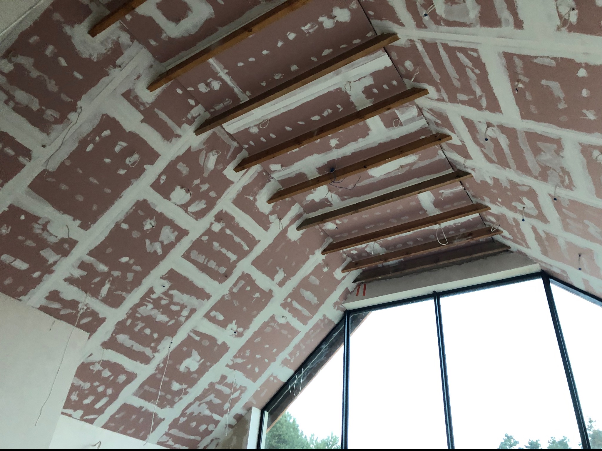 Skośny sufit poddasza z płyt karton-gips, wstępnie wyszpachlowany na łączeniach, widoczne drewniane belki konstrukcyjne i duże okno. Praca w trakcie realizacji.